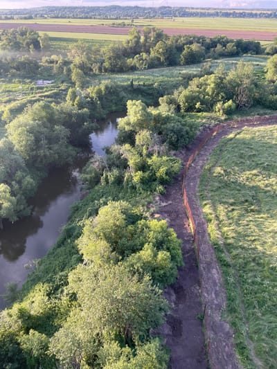 An aerial, wide-angle shot captures a landscape in Turovo, Russia, under a hazy sky. A dark, winding river dominates the left side of the frame, bordered by lush, green trees and dense foliage. A narrow, dirt path, partially fenced with orange posts, runs along the right bank of the river, leading into the distance. Beyond the trees and river, agricultural fields are visible, with some appearing to be cultivated in alternating strips of different colors, possibly representing different crops or stages of growth. The overall scene depicts a natural, rural setting with hints of human intervention in the form of the path and fence.