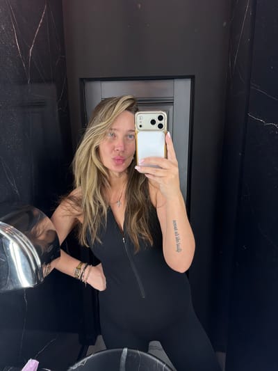 A woman with long, blonde hair is taking a selfie in a bathroom. She is wearing a black, zippered jumpsuit and several bracelets. A cross necklace is visible around her neck. She has a tattoo on her left forearm. The bathroom has dark, marbled walls and a silver hand dryer is visible on the left. The woman's facial expression is one of puckering her lips.