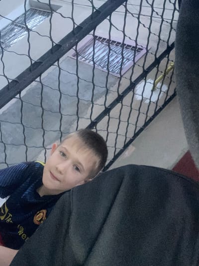 This media file is a selfie of a young boy. He is wearing a dark blue and yellow Manchester United jersey. The boy has fair skin, light brown hair, and blue eyes. He is smiling slightly and looking at the camera. The background shows a grey, textured wall with a metal fence in the foreground. Behind the fence, a building with windows can be seen. The lighting suggests it is daytime. The overall mood of the image is casual and happy.