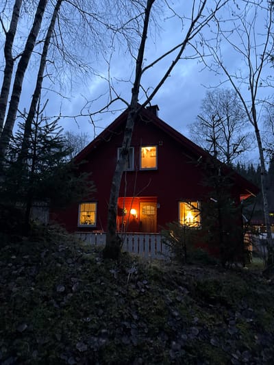 The image displays a red, two-story wooden house situated on an incline in Jessheim, Norway, during twilight hours. The sky is overcast, exhibiting blue and grey tones. The house features a dark gable roof with a chimney on the left side. Several windows are illuminated with warm, yellow interior light. A wooden front door with a small window pane is located on the ground floor, beside which an illuminated spherical white outdoor wall lamp is mounted. A white picket fence or railing spans the front of the house at ground level. A red-handled tool leans against the house wall near the door. The foreground consists of a sloping bank covered in dark green moss and scattered dark, fallen leaves. Bare deciduous trees with light bark are prominent in the foreground and around the house, their branches devoid of foliage. Smaller evergreen trees are also visible, some featuring small white elements. A darker forest line occupies the background. No individuals are present in the scene.