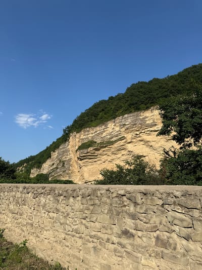 A high-angle, medium shot captures a natural landscape beneath a clear blue sky with a few wispy clouds. In the foreground, a rustic stone wall runs horizontally across the frame, constructed from irregular, light-colored stones with visible mortar. Green grass and weeds grow along the base of the wall. Beyond the wall, a steep, layered rock face dominates the middle ground, its warm, sandy-yellow tones contrasting with the dark green foliage that covers its upper slopes and the surrounding trees. The rock face exhibits distinct horizontal striations, indicating geological layers, with patches of greenery growing from crevices. The scene is located in the village of Khapil, Russia, suggesting a natural, mountainous or hilly terrain.