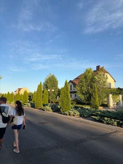 The image shows a street scene in what appears to be a residential area. Two people, a woman and a man, are walking on the asphalt road, which is bordered by a manicured hedge and several well-maintained cypress trees. Behind the hedge, a light-colored house with a brown roof is visible, partially obscured by trees. The woman is walking ahead of the man and is carrying a straw bag and a smartphone. She is wearing a white t-shirt and dark shorts. The man is behind her and is wearing a white t-shirt with a graphic print and dark shorts, with a black backpack slung over his shoulder. The sky is a clear, bright blue with a few wispy white clouds. The lighting suggests it is daytime, likely late afternoon or early evening, given the warm, soft light. The overall atmosphere is peaceful and casual, implying a leisurely stroll. No visible text is present in the image. The setting appears to be a quiet suburban street, possibly in Slobozhanske, Ukraine, as indicated by the location context.