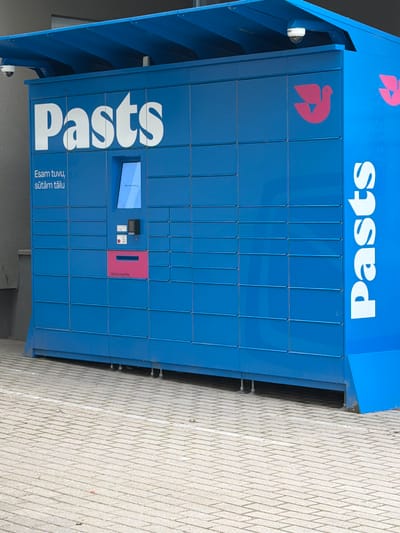 A bright blue parcel locker unit stands on a paved area in Upeslejas, Latvia. The unit has a large white logo that spells out "Pasts" across its front, and a smaller pink logo of a stylized bird is visible on the right side. Text in white, "Esam tuvu, sūtām tālu", is located on the lower left of the unit. A touchscreen interface and a slot for packages are integrated into the front of the locker system. The locker unit is positioned outdoors, with a gray building visible behind it and a tiled walkway in the foreground. A security camera is mounted on the overhang of the locker unit.