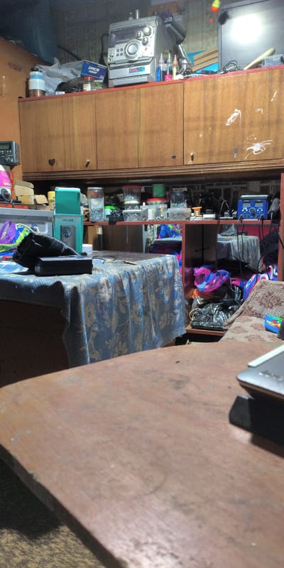 This image captures an indoor scene, likely a workshop or storage area, characterized by its cluttered and somewhat disheveled state. The dominant feature is a large wooden cabinet with multiple doors, filling much of the upper portion of the frame. The cabinet's surface is worn and shows signs of use, with some graffiti-like markings visible on the doors.
On top of the cabinet, various electronic devices and containers are piled. A prominent item is a silver stereo system with multiple dials and buttons. Next to it, several small bottles with colored liquids, possibly cleaning solutions or craft supplies, are arranged. A blue box labeled "DACE 4000P" is also visible, along with plastic bags and other miscellaneous items.
Below the cabinet, a red shelf unit is present, housing more containers filled with small objects, likely hardware like screws or nuts, as well as some tools and electronic components. A blue power supply unit with digital displays is also located on this shelf.
In the foreground, a table is covered with a blue and white floral patterned tablecloth. On this table, a black rectangular object, possibly a speaker or electronic device, and a green box with the label "JN-088" are placed. The table itself appears to be made of wood.
To the right of the table, a section of a patterned sofa or chair is visible, with some colorful plastic bags and black material placed on and around it. A small, colorful object, possibly a toy, is also present on the sofa.
The overall lighting is dim and artificial, suggesting an indoor environment with minimal natural light. The presence of tools, electronic components, and storage containers points towards a space used for repair, crafting, or hobbies. The location of Есипово, Russia, indicates a setting in a town known for its industrial history, which might explain the presence of such a workspace.