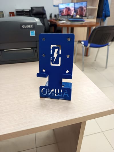 The image shows a blue metal phone stand with a lightning bolt symbol and stars cut out of it, positioned on a light wood-grain desk. The stand has the Cyrillic text "ОПЫША" cut out of its base. In the background, a black GoDEX printer is visible, along with a black office chair with blue accents. Further in the background, two computer monitors display images of rocky landscapes, and a blue jacket hangs on a shelf. The setting appears to be an office. There are no people visible in the image. The lighting suggests it is daytime, likely indoors with artificial light.