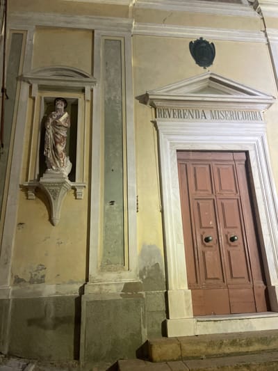 The image shows the facade of a building in Portoferraio, Italy, located at latitude 42.816 and longitude 10.332. The building features a yellow-painted wall with white trim and a large, wooden double door. The door is flanked by two white pillars and is topped with a marble archway.  Above the archway is a sign that reads "Reverenda Misericordia."  The building also has a small alcove on the left side, which contains a statue of a man holding a book. The alcove has a small, decorative pediment above it. The alcove's walls are painted a muted green and have white trim. There is a set of three stone steps leading up to the door. The steps are illuminated by artificial light,  casting shadows on the wall and steps. The surrounding environment is dark, creating a sense of mystery. The building appears to be old and well-maintained, with evidence of some wear and tear on its surface.  The scene is static, with no people or movement.