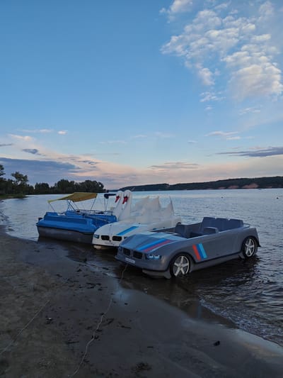 The image depicts a serene evening scene at a waterfront, likely Zelenodolsk, Russia, as suggested by the location context. The primary subjects are several pedal boats parked along a sandy shore. In the foreground, a grey pedal boat designed to resemble a car, complete with BMW-inspired racing stripes in red, white, and blue, is partially submerged in the calm water. Adjacent to it are two white pedal boats shaped like swans, also waiting to be used. Behind these, a blue pedal boat with a yellow canopy is moored.
The setting is a tranquil lake or river, with a tree-lined shore visible in the distance across the water. The sky above is a gradient of soft blues and pinks, indicating either sunrise or sunset, with scattered white clouds adding texture. The overall mood is peaceful and inviting, suggesting a leisurely atmosphere.
There are no people visible in the image, implying the boats are either waiting for customers or have been left by their users. The water is gently lapping at the shore and the boats, reflecting the soft light of the sky. The sandy beach is wet in places from the water's edge, with some debris and a few thin ropes extending from the boats onto the sand, possibly for mooring.
Visible text on the grey car-shaped boat, above the front wheel, reads "kalamaranfix.ru". Similar text is present on the white swan-shaped boat. The time of day appears to be twilight, given the soft lighting and pastel colors in the sky. The weather seems calm and pleasant, conducive to a day out on the water.