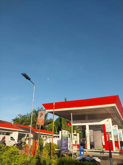 The image depicts an outdoor scene at a Pertamina gas station in Macege, Indonesia. The sky is a clear, bright blue, suggesting it is daytime, likely morning or afternoon, with a visible moon. The dominant structure is the red and white gas station canopy, with its distinctive angled roof.

Several gas pumps are visible, some displaying the "PERTAMAX TURBO" and "PERTALITE" branding, indicating the types of fuel offered. One pump has a blue cylindrical component attached. There are also signs with informational text and symbols, including one with an orange circle and a diagonal line over a "P" symbol, indicating no parking. Another sign below it reads "DILARANG PARKIR DI AREA INI," which translates to "NO PARKING IN THIS AREA." Other signs are in Indonesian and appear to be informational or promotional.

In the foreground, a modern street light with an LED panel is visible, angled upwards. To the right, lush green foliage surrounds the gas station. A white van is parked near the pumps, and a person wearing a red helmet is interacting with it. A motorcycle is also parked nearby.

The overall atmosphere is functional and everyday, capturing a typical scene at a petrol station. The bright sunlight and clear sky contribute to a sense of an ordinary day.