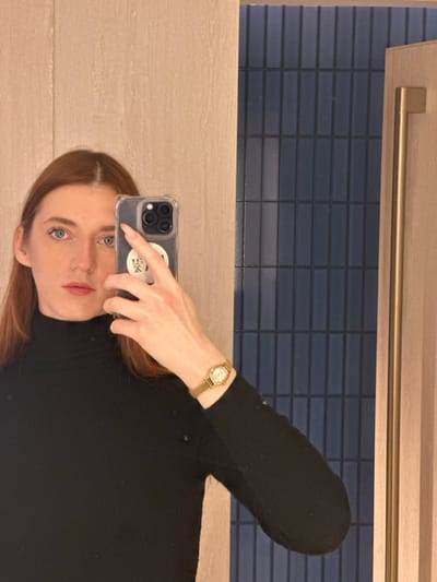 A mirrored selfie of a woman in a black long-sleeved shirt is angled in the image. She holds a phone up to the mirror with her left hand. A gold watch is visible on her left wrist. Her red hair is parted in the middle. In the background is a tan countertop, dark blue brick tiling, and a gold towel rack.