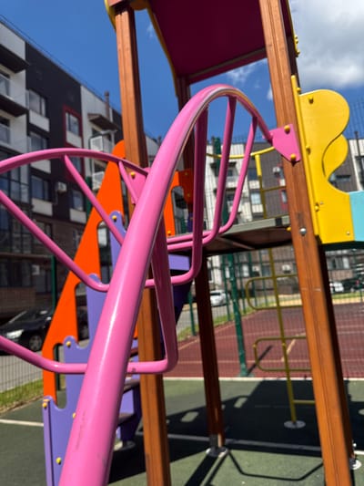 A close-up, low-angle shot of a brightly colored playground structure, with pink, orange, and yellow elements prominently featured. The playground is situated in a modern urban setting, with contemporary apartment buildings visible in the background. The sky is clear and blue with scattered white clouds. The ground surface appears to be a mix of green rubberized material and red track-like flooring, common in public play areas. The image captures a sunny day, with shadows cast by the playground equipment. The location is identified as Vyazniki, Russia.