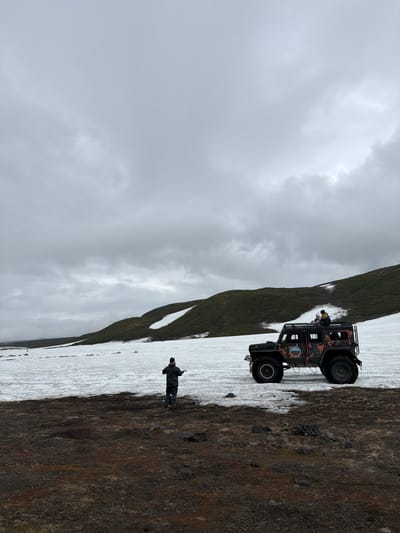 The image depicts a bleak, overcast day in a mountainous, possibly volcanic, landscape near Petropavlovsk-Kamchatskiy, Russia. The foreground consists of dark, rocky, and sparsely vegetated terrain. A wide expanse of melting snow covers the middle ground, with patches of dark earth visible through the snow. In the mid-ground, a person wearing a dark jacket and cap is standing with their back to the camera, facing the snowy landscape and a large, dark-colored off-road vehicle. The vehicle is heavily modified with large tires and a roof rack. Two individuals are seated on the roof of the vehicle, one appearing to be a child. In the background, rolling hills covered in dark green vegetation rise steeply, with streaks of snow visible on their slopes. The sky is a uniform, heavy gray, suggesting cold weather or imminent precipitation. The overall scene is desolate and rugged, characteristic of a remote wilderness area.