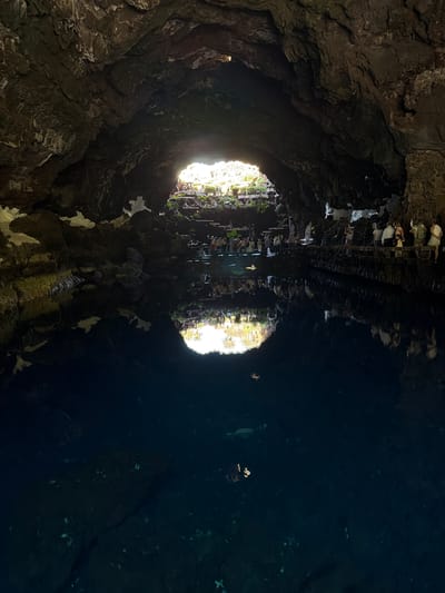 This striking image depicts the interior of a dark, rocky cavern or lava tube, featuring a large, still body of water acting as a prominent reflective surface. The perspective is from within the cavern, looking towards two bright, circular openings in the distance that allow natural light to stream in.

On the left side of the frame, the rough, textured walls and ceiling of the cavern are visible, characterized by dark brown, gray, and black rock formations. These formations show intricate patterns and crevices, illuminated subtly by the distant light.

The right side of the image is dominated by the calm, deep blue-black water, which perfectly mirrors the scene above and around it. The reflections in the water show the bright openings, appearing as two circular sources of light, and the inverted images of the rocky structures. Most notably, the reflections clearly show numerous silhouettes of people, appearing upright in the water. These individuals are likely standing or seated along a pathway or viewing area opposite the water's edge, outside the immediate frame of the actual scene, with only their reflections being fully captured. Faint green hues from distant vegetation are also discernable around the reflected light sources, suggesting an outdoor area beyond the cavern openings.

The time of day appears to be during daylight hours, given the bright natural light. The strong contrast between the dark cavern and the intensely lit openings creates a dramatic and artistic visual. The setting, identified as Haría, Spain, strongly suggests this is a converted volcanic cave system, possibly Jameos del Agua, designed for public visitation. There is no visible text in the image.