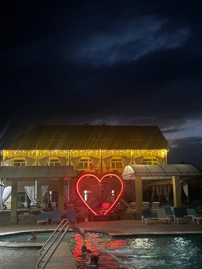 The image depicts a nighttime scene at an outdoor swimming pool facility, possibly in the city of Yaroslavskaya, Russia. In the foreground, a swimming pool is illuminated by a large, red neon heart-shaped sculpture. Several people are visible in the pool, some partially submerged and engaged in leisure activities. To the right of the pool, there are lounge chairs and covered seating areas. In the background, a building with a yellow-tiled roof is adorned with a string of small yellow lights along its eaves, and there are illuminated windows. The sky above is dark with visible cloud formations, suggesting evening or night. The overall atmosphere suggests a recreational area operational after dark.