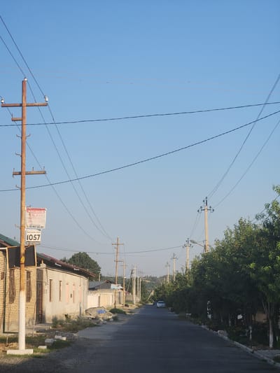 This image depicts a residential street in Xidirsho, Uzbekistan, under a clear blue sky. A prominent rusty metal utility pole stands on the left side of the road, adorned with a sign that reads "ABU AYUB GILAM MEBEL YUVISH" and a phone number, followed by "1057 bekijaan taxi". Adjacent to the pole is a building constructed with light-colored brick. Across the street, a row of trees lines the pavement, some with whitewashed trunks. Further down the street, the road curves gently, with more utility poles and trees visible. A white car is parked on the right side of the road, and another vehicle is moving away in the distance. The overall impression is one of a quiet, sunny day in a semi-rural or suburban setting.