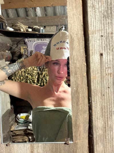 In a dimly lit, rustic room that appears to be part of a sauna or bathhouse in Maksimovo, Russia, a woman stands in front of a mirror. She is wearing a green towel wrapped around her body and a grey and white felt hat adorned with Cyrillic lettering. The hat is positioned on her head and she is holding it with her left hand. The woman has tattoos on her left arm and shoulder. The background of the image shows a cluttered environment with wooden walls, shelves filled with various objects including coils of wire, buckets, and what appear to be bundles of dried herbs. A rough wooden structure frames the right side of the image, creating a strong vertical element. The overall impression is one of a simple, utilitarian space.