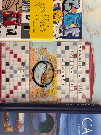 The image depicts a visually complex arrangement of items, including a map, Scrabble tiles, graffiti art, and books. The Scrabble board features the words "MONKEY FOLLOW THE FEET". The map underneath shows parts of Europe, North Africa, and the Middle East, with cities like Lisbon, Morocco, Algeria, Egypt, and Syria labeled. Several colorful graffiti art pieces are visible at the top of the image, overlaid on other abstract designs. Below the map and Scrabble board, a dark blue book with the word "CAMINOEN" is present, and further down, another book with the word "CA Pilgrimsfæ" is partially visible. A black cord with a small, colorful pendant is draped over the map, near a Scrabble tile labeled "E". The overall scene appears to be a still life composition, potentially an art installation or a meticulously arranged display, and there is no indication of a specific location within Malmö, Sweden.