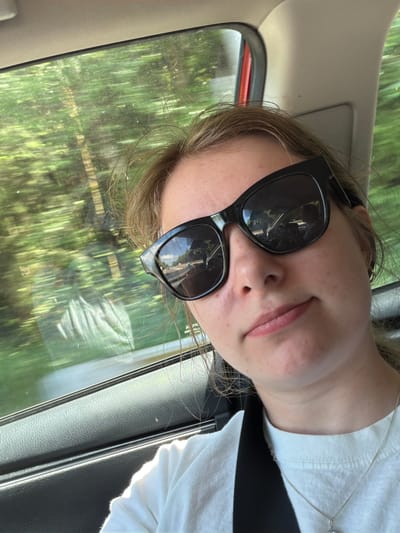 A young Caucasian female is taking a selfie inside a vehicle. She is wearing black sunglasses, a white t-shirt, and a silver necklace. The background shows a blurred view of trees and sky through the car window, indicating movement. The interior of the car is visible, including the car door and part of the headliner. The image appears to be taken during the daytime, likely on a sunny day. The car is likely traveling on a road within or near Seaside, United States, as suggested by the presence of trees and a highway visible in the reflection of her sunglasses.
