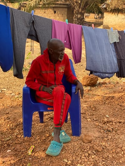 The image captures an outdoor scene in the city of Nev, Nigeria. A single elderly Black male, bald with a white beard, is seated on a blue plastic "OCEAN" chair. He is dressed in a red tracksuit featuring green stripes along the sleeves and legs, and the jacket has a zippered front with a light-colored animal graphic, possibly a tiger, on the left chest. He wears teal slip-on clogs. His right leg is crossed over his left, and his hands rest on his lap. His gaze is directed to the right, out of the frame. Behind the individual, a clothesline is suspended, from which various items of clothing—including blue, grey, and purple trousers, a patterned blue fabric, a light green shirt, and a dark patterned garment—are hung with blue and green plastic clothespins. The ground is an expanse of reddish-brown soil mixed with gravel and small stones. In the background, an earthen-colored building, constructed from what appears to be mud or similar material, is partially visible behind a large palm tree, with additional structures discernible in the distance. A small yellow wrapper is present on the ground near the man's feet, and a reddish-brown rock sits to his right.