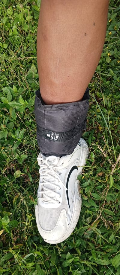 A person's lower right leg and foot are positioned vertically on a verdant, overgrown patch of grass and broadleaf plants in Weddagala, Sri Lanka. The leg displays bare, medium-brown skin with visible pores, a light tan line around the ankle, and several small blemishes or insect bites on the calf. A black fabric ankle weight, marked with a white "SUZUKI 1 Kg" label, is secured around the ankle. Below the ankle weight, a white athletic shoe with black accents and white laces is worn. The shoe's surface appears slightly worn and soiled, consistent with outdoor use. The surrounding ground is densely covered with various green foliage, including slender blades of grass and wider leaves, with some small yellow flowers interspersed throughout.