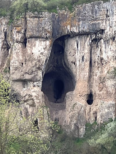 The image shows a rocky cliff face with a large, irregular cave entrance in the center. There is a smaller hole higher up on the cliff face to the right of the cave entrance. The cliff face appears to be made of light gray and brown rock with layered formations. Vegetation is visible along the edges of the cliff, consisting of green foliage and trees. The large cave entrance is dark, suggesting it extends deep into the rock. The image is taken in Barkach, Bulgaria.