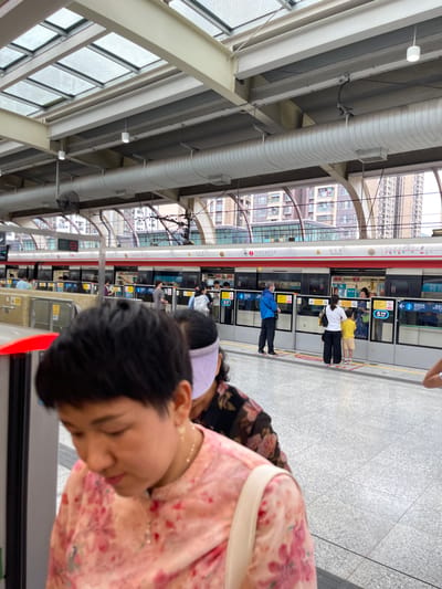 This media file depicts a scene at a subway station in Longhua District, China. The primary subjects are several people waiting on the platform for a subway train. The setting is an indoor subway station with a modern architectural design, characterized by exposed structural beams, ribbed ceilings, and large windows allowing natural light. A subway train with red and white livery is visible at the platform, with its doors open.
The people appear to be commuters, with some standing and others looking towards the train. One individual in a blue jacket is visible boarding or disembarking. The overall atmosphere suggests a typical day at a busy transit hub. Visible text on the train includes station names and route information in Chinese characters, such as "白石龙 Baishilong" and "4号线往福田口岸" (Line 4 towards Futian Port). The time of day is unclear, but the lighting suggests it is daytime. There are no strong indicators of specific weather conditions. The image is captured from a low angle, focusing on the people and the train.