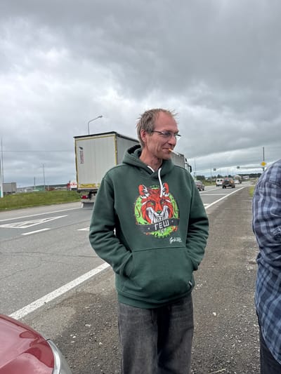 An overcast day at an exit or entrance to Alnashi, Russia, on a roadside. A Caucasian man with thinning blond hair, wearing glasses and a dark green hooded sweatshirt with a graphic of a red fox and the text "ГЕШ Gesh Style", stands with his hands in his pockets, smoking a cigarette. He is on the side of a road with multiple lanes, a white line marking the edge of the road, and an off-ramp visible in the distance. A large white semi-trailer truck, labeled "SCHMITZ CARGOBULL", is parked on the side of the road behind him. Several other vehicles are visible on the road, including cars and a cement mixer truck. The sky is cloudy and gray. Another person, wearing a blue and gray plaid shirt, is standing to the right of the frame, partially visible. A portion of a red car is visible in the lower left corner of the image.