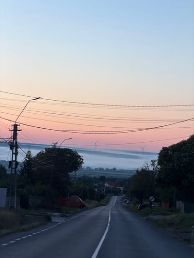 The image depicts a rural landscape at dawn, with a road leading into the distance towards a valley filled with mist. Several wind turbines are visible in the background, indicating the presence of a wind farm. The sky displays a gradient of colors, transitioning from pale blue at the top to soft pink and orange hues near the horizon, characteristic of sunrise. Power lines and utility poles are prominent in the foreground, crossing the scene horizontally. A few trees and houses are scattered throughout the landscape, suggesting a small village or rural settlement. The road is asphalted and has a white line marking the center. An orange van is parked on the side of the road on the left, partially obscured by vegetation. The overall atmosphere is serene and tranquil, characteristic of a quiet morning in the countryside. The location is believed to be near Pașcani, Romania, given the visual elements common to such areas.