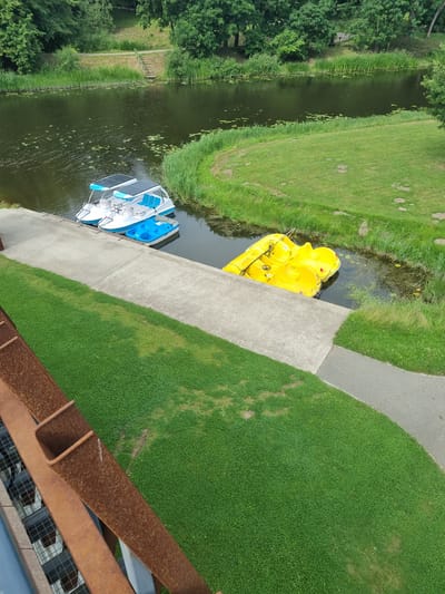 The image shows a top-down view of a canal or narrow river with two pedal boats docked at the edge. One boat is a bright yellow, duck-shaped pedal boat, and the other is a white and blue pedal boat with solar panels on top. The canal is surrounded by lush green grass and trees. A paved pathway runs alongside the grass, bordering the canal. The scene appears to be a park or recreational area. The water is calm and reflects the surrounding greenery. There is no visible text in the image. The lighting suggests it is daytime, with the sun casting shadows on the grass, indicating a sunny day. The setting is a peaceful outdoor environment, likely in a suburban or rural area given the presence of natural elements and recreational facilities. The image does not depict any people or activity, focusing solely on the static scene of the boats and the landscape. The rusted metal railing in the foreground suggests the vantage point is from a bridge or elevated structure.