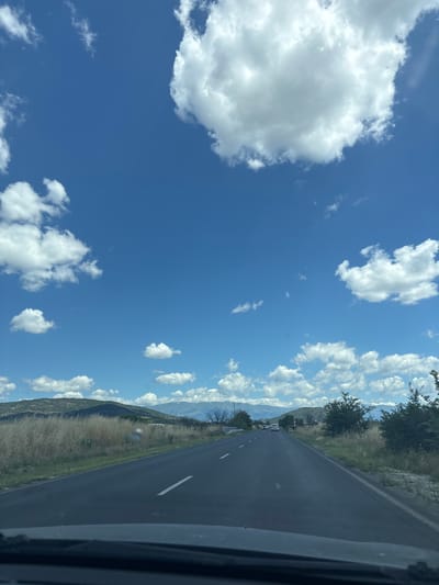 The image shows a clear blue sky filled with scattered white clouds. A dark asphalt road, marked with white lines, stretches into the distance, appearing to be a highway or a major road. The roadside is lined with dry, golden grass and scattered trees, with rolling hills and mountains visible in the background. Some of the distant mountains appear to have snow-capped peaks. A few cars are visible on the road ahead. The foreground shows the dashboard and windshield of a vehicle, indicating the image was taken from inside a car. The scene appears to be in a rural or semi-rural area under a bright, sunny sky.