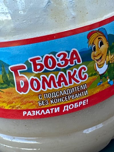 The image shows a close-up of a label on a food product. The label is predominantly red, blue, and yellow, with white text and a cartoon character. The text appears to be in Cyrillic script and reads "БОЗА" and "БОМАКС". Below this, there is smaller text that translates to "WITH SWEETENERS WITHOUT PRESERVATIVES" and "SHAKE WELL!". The cartoon character is a smiling boy with blond hair, wearing a red and blue cap, a white tank top, green shorts, and blue shoes. He is positioned to the right of the text. The background of the label depicts a landscape with hills and trees under a blue sky. The product itself appears to be a creamy, pale substance contained in a clear jar or bottle, which is slightly out of focus. The lighting suggests an outdoor setting. There is no visible indication of the city of Chernomorets, Bulgaria in the image.