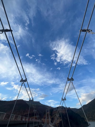 The image presents a view looking up from a suspension bridge towards a partly cloudy sky. Metal suspension cables stretch upwards from the lower left and right corners towards the center of the frame, supporting the bridge. The sky is a mix of blue and white clouds. On the right side of the image, a mountain slope is visible, its vegetation a mix of green and brown. The structure of the bridge itself, with railings, can be seen on the right side as well. The location is Ait Mazigh, Morocco. The bridge spans a valley or gorge. There are no people visible.