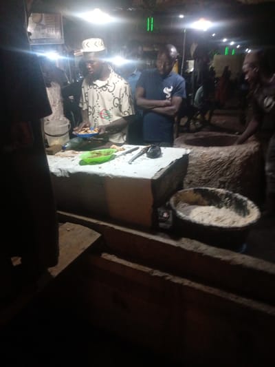 In the city of Tundun Wada South, Nigeria, a group of individuals are gathered around a food stall at night. The stall is illuminated by bright overhead lights, creating a somewhat hazy atmosphere. In the foreground, a man wearing a white cap and a patterned shirt is serving food from a blue plate, holding utensils. To his right, another man in a dark blue t-shirt stands with his arms crossed, observing.
The food stall itself is a makeshift setup with a white tablecloth covering a surface where food preparation appears to be taking place. Various bowls and containers are visible, including a green bowl and a large, rustic-looking basin filled with what appears to be a dry ingredient, possibly flour or grains. In the background, other figures are present, some blurred due to motion or low light, suggesting a bustling market or street food scene. The ground beneath the stall is uneven and appears to be made of packed earth or concrete. The overall environment suggests an outdoor setting, likely a market or a roadside vendor, characterized by its nighttime activity and vibrant, if somewhat dim, illumination.