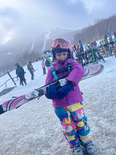 A young child, likely a girl, stands on a snow-covered surface at a ski resort, looking directly at the camera with a neutral expression. She is dressed in full ski gear, including a bright purple ski jacket, vibrant multi-colored ski pants with a geometric pattern, a purple helmet, and goggles with an orange tint. She wears blue gloves and has skis, which appear to be Rossignol brand, attached to her feet.
In the background, a long rack filled with numerous skis and snowboards is visible. Beyond the rack, other skiers and snowboarders can be seen on the snowy slope, some appearing to be waiting in a line. The setting suggests a busy day at a ski area. The weather is overcast, with a hazy, cloudy sky dominating the upper left portion of the image. Distant mountain peaks or tree lines, also covered in snow, are faintly visible through the mist. The ground is a mix of packed snow and possibly some icy patches, typical of a well-used ski area. The overall scene depicts a winter day at a ski resort in Jay, United States. The text "PROD" is partially visible on the child's goggles, and "ROSSIGNOL" is visible on the ski bindings.