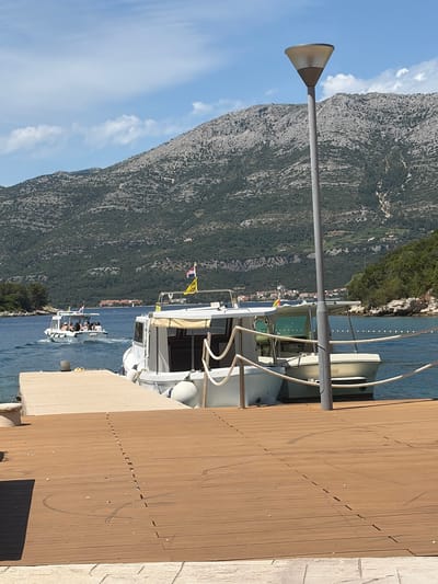 The image depicts a sunlit coastal scene in Grad Korčula, Croatia. In the foreground, a brown wooden deck occupies the lower half of the frame, constructed from horizontally laid planks with a central vertical seam. Faint, dark, curved scuff marks are visible on this surface. To the left, a light-colored concrete pier extends diagonally into the water, on which a pair of yellow flip-flops rests. A grey, cylindrical lamppost with a conical, beige-lined fixture stands vertically, slightly right of the frame's center.
In the midground, the calm blue water of a bay is visible. Two white motorboats are docked alongside the concrete pier on the right, secured by ropes, one of which is partially draped near the lamppost. The closer of these boats features beige canvas coverings. A line of white spherical buoys extends across the water further out, delimiting an area. To the left, a smaller white passenger boat, carrying several individuals, moves away from the viewer, creating a wake. This boat flies a Croatian flag and a yellow flag with partially visible black text.
The background is dominated by large, rugged mountains characterized by grey rock and sparse green vegetation. The sky above is clear blue with scattered white, wispy clouds. Along the distant shoreline, at the base of the mountains, a cluster of buildings with orange roofs is discernible, indicating a settlement within the broader Grad Korčula area.