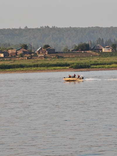 A motorboat with three occupants is traversing a wide, calm river. The boat is light-colored and appears to be moving at a moderate speed, leaving a wake behind it. The occupants are wearing orange life vests. In the background, a distant shoreline features a cluster of rustic wooden houses and buildings, suggesting a rural or semi-rural settlement. Beyond the settlement, a dense forest covers a rolling hillside. The sky above is overcast with a soft, hazy light. The overall scene depicts a tranquil day on a river in a natural setting with evidence of human habitation.