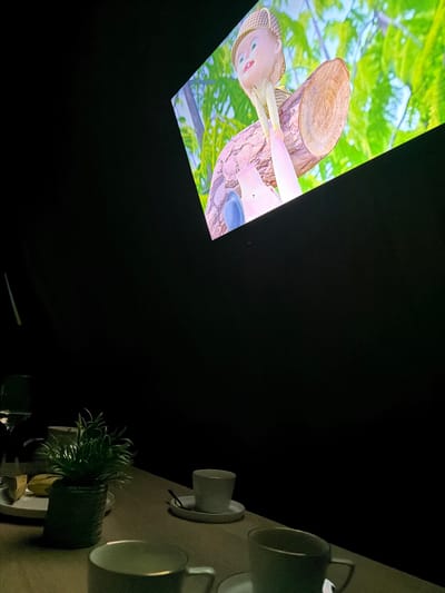 In Sharovechka, Ukraine, a dimly lit room features a projection of a cartoon character, possibly Pinocchio, sitting on a log amidst vibrant green foliage. The projection is cast onto a dark surface, angled upwards. In the foreground, a wooden table is set with two white teacups and saucers, and a small potted plant. To the left, a plate holds a piece of bread or cake, next to a wine glass. The overall scene suggests a cozy, indoor viewing experience.