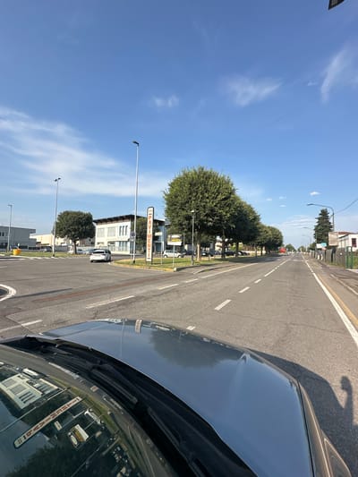 The image shows a street scene in Rudiano, Italy, viewed from the perspective of someone inside a car. The car's hood and windshield are visible in the foreground, reflecting the surrounding environment. The street is paved with asphalt and marked with white lines indicating traffic lanes. On the left side of the street, there is a roundabout with a yellow bollard. Further down the street, there are buildings, trees, and streetlights. Several cars are parked or driving on the street. A tall vertical sign on the right side of the road reads "PAVIMENTI" with an arrow pointing to the right, indicating flooring or pavement services. The sky is blue with scattered white clouds. The overall scene depicts a typical urban or suburban environment during the daytime.