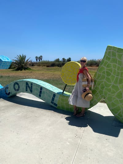 A woman with blonde hair and sunglasses, wearing a white sundress, a red scarf, and sandals, poses for a photo in front of a large, mosaic-tiled art installation in Conil De La Frontera, Spain. The art piece spells out "CONIL" and features vibrant blue, green, and yellow tiles. The woman is holding a straw bag and a straw hat. The background consists of a grassy field, palm trees, and a clear blue sky. The ground is paved with concrete.