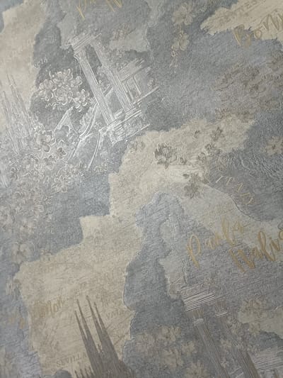 The image displays a close-up view of wallpaper with a map-like design featuring faded text and architectural elements. The wallpaper has a textured appearance, with areas of light and dark gray, and lighter beige or cream-colored sections. Visible text includes "MADRID," "VALENCIA," "MILANO," "ITALY," "ROME," and what appears to be "SEVILLA." There are also stylized drawings of classical columns and floral motifs. In the lower right section, there's a golden script that reads "Parigi Italia." The wallpaper design evokes a travel or European city theme. The image does not depict any people, actions, or interactions. It is a still image of a decorative surface. The location in the city of Giaginskaya, Russia, is not evident from the image itself.