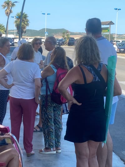 The image shows a group of people standing on a paved area with parked cars and palm trees in the background. The location is identified as Conil de la Frontera, Spain, with a sign indicating "Río Salado" visible.
In the foreground, several individuals are captured from the waist down. On the left, a person is seated in a chair, with their legs visible. To their right, a woman is standing, wearing burgundy pants and a white t-shirt. Next to her, another woman stands with her back to the camera, wearing a light blue tank top and patterned pants, with a red backpack. Further to the right, a woman in a black tank top and black skirt is standing with her hands on her hips. She is holding a green object, possibly a rolled-up umbrella or mat, in her right hand. A man is partially visible to her right, wearing a light blue shirt and light blue shorts.
In the middle ground, several other people are visible, including a man and two women interacting. One woman is wearing sunglasses and a white t-shirt. The background features parked cars, including black and white vehicles, and a hilly landscape with vegetation under a clear blue sky. Streetlights are visible in the background. The overall scene appears to be a public area in a sunny, outdoor environment.