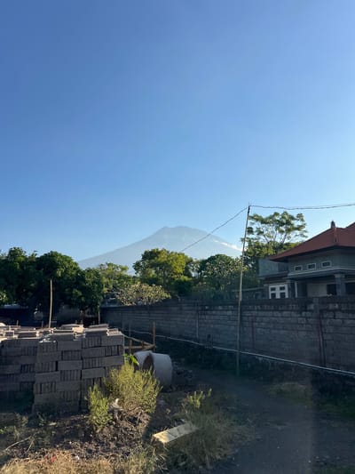 The image depicts an outdoor scene in Kubu, Indonesia. The foreground on the right shows a pile of gray bricks, some dry grass, and disturbed earth, suggesting a construction or renovation site. Behind the bricks, there is a large concrete pipe, possibly for drainage or construction. Further into the scene, a low brick wall separates the construction area from a building with a red tiled roof. This building appears to be made of dark stone or brick and has several windows. Beyond the building, lush green trees are visible, with a majestic mountain peak rising in the distance, partially obscured by haze. The sky is a clear, bright blue, indicating a sunny day, likely in the late morning or early afternoon given the sunlight's angle. There are power lines running across the scene, typical of a semi-urban or rural setting. The overall impression is one of ongoing construction set against a backdrop of natural beauty, characteristic of a tropical or subtropical location like Indonesia. There is no text visible in the image.