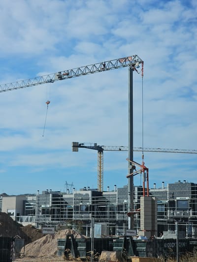 The image depicts a construction site in Cuarte de Huerva, Spain, under a partly cloudy blue sky. The foreground shows a chain-link fence bordering piles of dirt and sand, with several dark-colored dumpsters visible. In the midground, a multi-story residential building is under construction, covered in scaffolding and grey cladding. The building features numerous windows, and the word "WERU" is visible on some of them. Signs with "PRIONSA" are also present on the building.
Dominating the scene are several construction cranes. A large, latticed tower crane extends across the upper portion of the image from left to right. A smaller tower crane, predominantly yellow and grey, is positioned further back to the right. A third, more central crane consists of a tall grey pole with a red and orange jib mechanism attached near its top, holding a stack of concrete blocks. A hook and cable hang from the jib. The overall impression is one of ongoing construction and development in a suburban area.