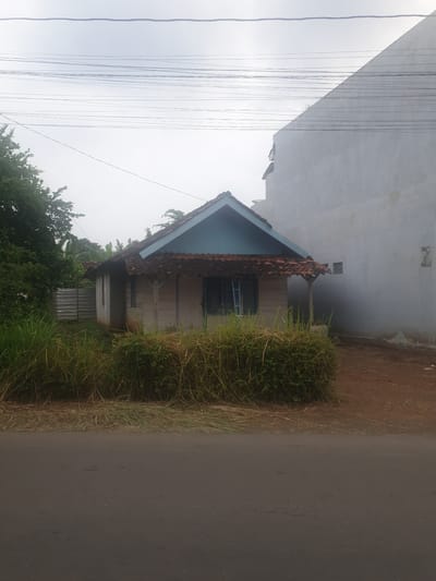 The image depicts a street scene in Proyonangg, Indonesia, characterized by a modest house on the left and a paved road on the right. The house appears to be constructed with concrete blocks and has a tiled roof. A portion of the house has a light blue painted facade under the eaves. In front of the house, there is a dense growth of tall grass and bushes, with some trees visible further in the background. Overhead, several dark wires are suspended, suggesting the presence of electrical or telecommunication lines. The sky is overcast, indicating a cloudy or possibly rainy day. The time of day is indeterminate due to the lighting conditions. The scene is static, with no people or specific activities evident. The overall impression is one of a quiet, everyday residential area.