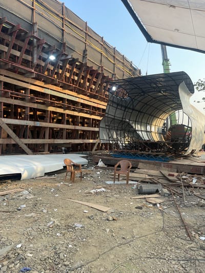 The image depicts a construction site in Mira-Bhayander, India. A large, curved metal structure, partially covered in white material, dominates the scene. It is supported by a complex wooden scaffolding system, some of which is covered with dark sheeting. A crane is visible in the background, suggesting ongoing construction. Two tan-colored plastic chairs are located on the ground near the base of the structure. The ground is covered in dirt, gravel, and construction debris. No people are visible.