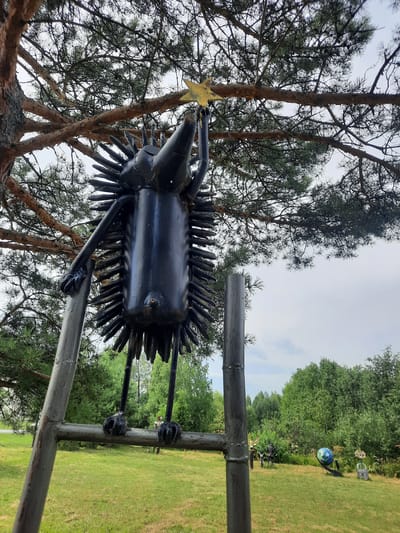 A black metal sculpture of a hedgehog is depicted outdoors, reaching for a yellow star above it. The sculpture is positioned on a ladder made of metal poles, with its feet resting on a horizontal bar of the ladder. The hedgehog sculpture is intricate, with spiky details resembling quills and a long snout. Its left arm is extended upwards towards the star.
The background shows a grassy field with trees and a cloudy sky. In the distance, other metal sculptures are visible, including a globe and a padlock. The scene appears to be in a park or an outdoor art exhibition. The specific location is identified as the city of Zelenogorsk, Russia. There are no people clearly visible interacting with the main sculpture, though there might be some figures in the far distance with other sculptures. The environment is natural, with a focus on the sculptures as the primary subject.
