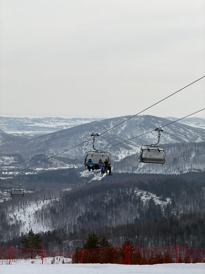 This image depicts a winter scene at a mountainous ski resort in Novoabzakovo, Russia. The sky is overcast and light grey, covering the upper half of the frame. Below the sky, a vast expanse of snow-covered mountains and valleys extends into the distance, with visible patches of bare deciduous and evergreen trees. A ski lift system dominates the mid-ground, featuring cables stretching diagonally. One four-person chairlift, positioned centrally and moving from left to right, carries three individuals. These individuals are dressed in winter ski jackets (blue, light blue/purple, and yellow/black from left to right) and protective headwear. The person on the far right has skis attached to their boots, dangling below the chair. Another empty chairlift is visible to the right on the same cable, and a third empty chair is partially visible further left and lower down. The immediate foreground consists of a snow-covered slope, edged by an orange safety net supported by red poles, with several evergreen trees positioned behind the net.