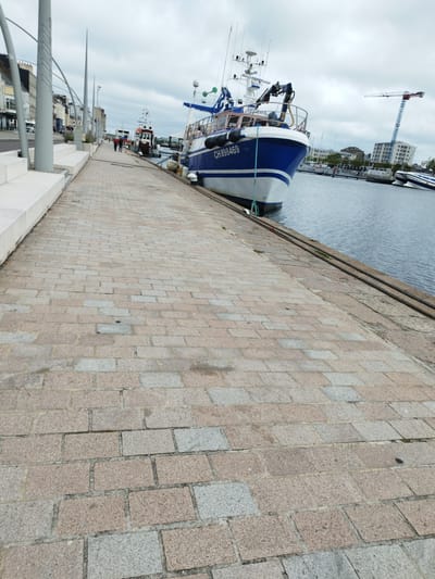 A quay in Cherbourg-en-Cotentin, France, is depicted under a cloudy sky. The foreground features a cobblestone path, while a blue and white fishing boat, identified by the registration "CH898469," is docked to the right. Further along the quay, other boats and buildings line the waterfront, with a construction crane visible in the background. Several individuals are walking along the quay.