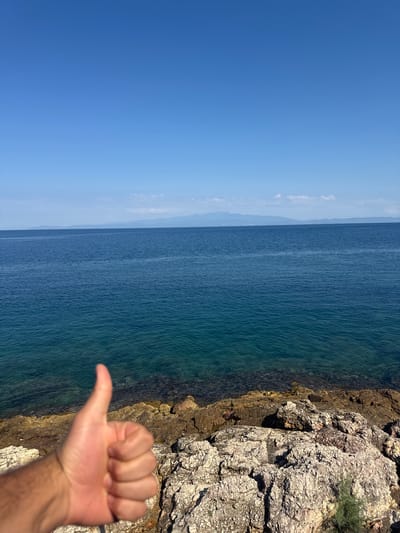 A human hand with a thumb raised in a "thumbs up" gesture is in the foreground, obscuring the lower left portion of the image. The hand is positioned over a rocky shoreline made up of light-colored, weathered rocks. The sea, with clear, turquoise water in the shallows and a deeper blue further out, stretches to the horizon. In the distance, a hazy, mountainous landmass is visible under a clear blue sky with a few wispy clouds. This scene is located on the coast of Skala Marion, Greece.