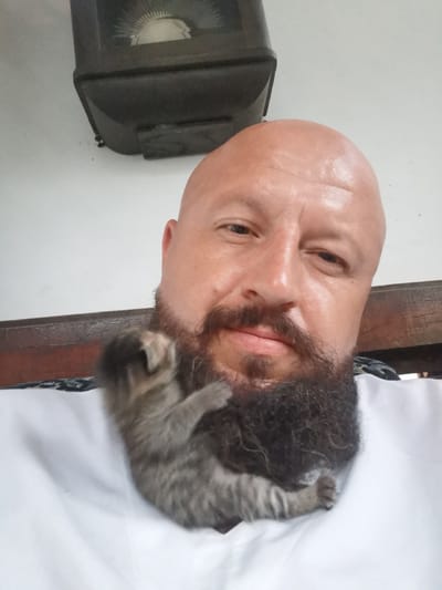 A close-up, low-angle selfie shows a bald man with a full beard, wearing a white shirt. He is holding a small, grey tabby kitten that is clinging to his beard. The man is looking towards the camera, with his head tilted slightly. A dark, rectangular object with a circular dial is mounted on a white wall behind him. To the right, a wooden beam runs horizontally. The background is a plain white wall. The scene is indoors. The image does not provide any information about the location in the city of Semypolky, Ukraine.