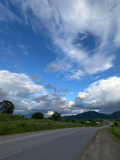 A paved road winds through a grassy landscape with scattered trees under a partly cloudy sky. In the distance, a village is nestled at the base of a tree-covered mountain range. Two cars are visible on the road in the middle ground. The scene appears to be a rural setting, possibly in or near Teploozersk, Russia.